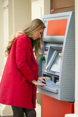 Fototapeta na wymiar Young blonde woman in a red coat withdraw cash from an ATM in th