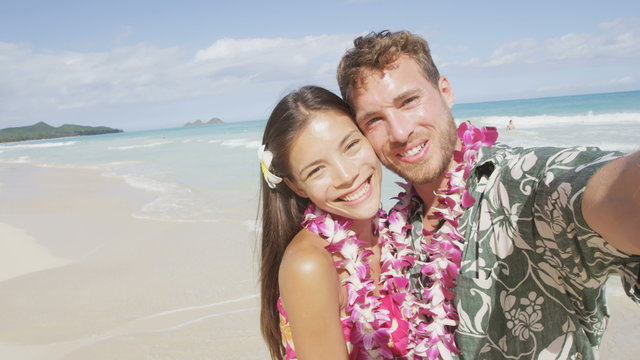 Selfie couple in love on hawaii beach taking photo self portrait. Young woman and man in love using mobile cell smart phone on Hawaii beach vacation in Hawaiian clothing wearing flower lei. RED EPIC.