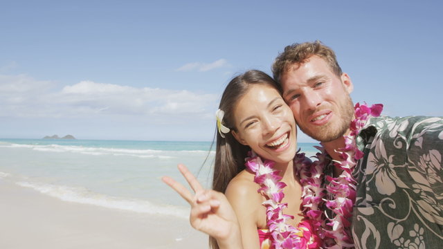 Couple on beach taking selfie photo on Hawaii with smart phone.  Young woman and man in love using smartphone on beach vacations in Hawaiian clothing wearing Aloha shirt dress and flower lei. RED EPIC