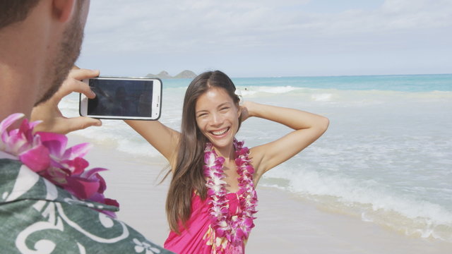 Man taking pictures with smart phone of woman on beach on Hawaii. Young couple having fun living happy lifestyle on Hawaiian beach holiday vacation travel. Man using smartphone for photo. RED EPIC.