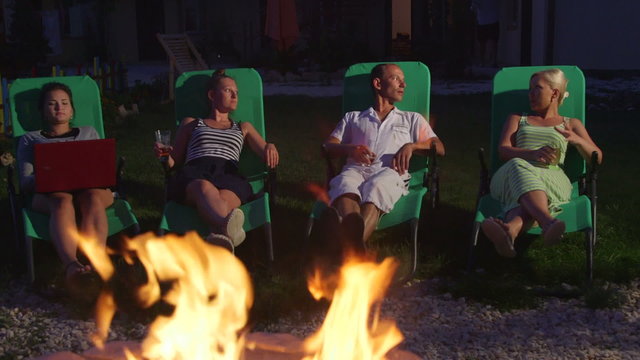 Family With Friends Relaxing On Grass Lawn In Backyard Near Stone Fire Pit