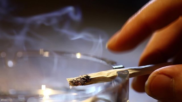 Color footage of a cigarette, burning in an ashtray and a hand.