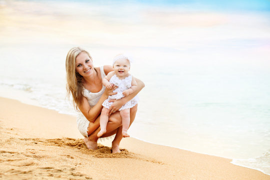 Happy Mother And Baby Girl Sitting On Beach Near Sea