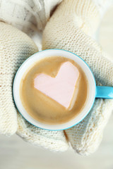 Female hands in warm mittens holding cup of hot cappuccino with heart marshmallow, close up
