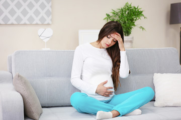 Pregnant woman with headache sitting on sofa in the room