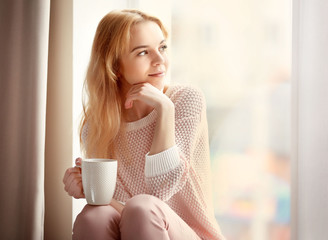 Blonde beautiful  girl  sitting by the window with cup of tea or coffee in her hands on the light background