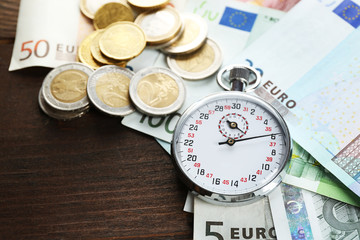 Stopwatch with euro banknotes and coins on wooden background. Time is money concept