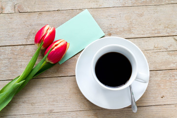 Red tulips with a cup of coffee and a message card on a wooden table, top view.