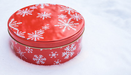 Christmas gift container.  Red, round cookie & baked goods tin container decorated in red with white snowflake print pattern, sitting in natural snow. - 105845955