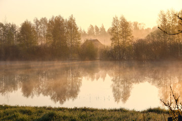 Foggy landscape: early morning on the river in the spring, the silhouette of  wooden house over the water. Village, countryside. Fog. Sun.