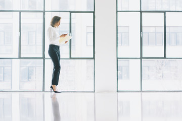 Fototapeta na wymiar Photo business woman wearing modern suit, looking mobile phone and holding papers in hands. Open space loft office. Panoramic windows background. Horizontal mockup.