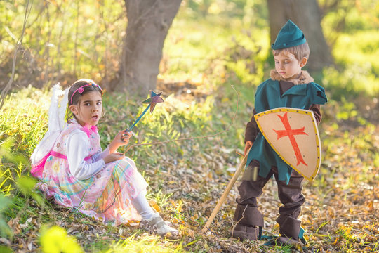 Portrait of a cute little girl dressed up as a fairy sitting in a magical forest and playing with a boy dressed up as a knight