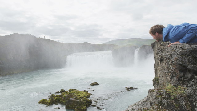 Tourist enjoying view of waterfall Godafoss on Iceland. Man lying on cliff edge looking down on travel visiting tourist attractions and landmarks in Icelandic nature on Ring Road. RED EPIC.