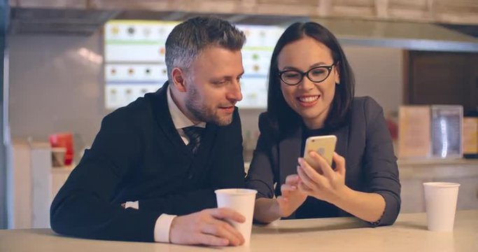 Asian businesswoman showing her male colleague something on her smartphone while sitting in the cafe, they talking and smiling