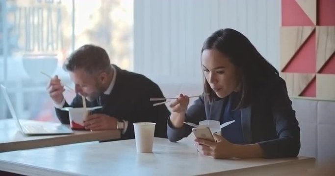 Asian businesswoman using her smartphone while having lunch in noodles bar, mid-adult businessman sitting next to her, eating and typing on his laptop