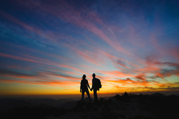 Fototapeta na wymiar Silhouettes of two hikers with backpacks enjoying sunset view from top of a mountain. Travel concept