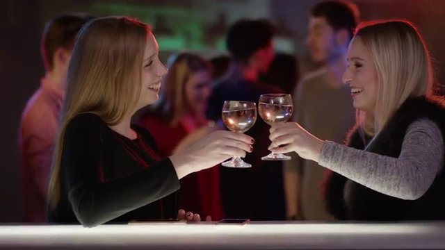 two attractive girls at bar in club talking and raising their glasses