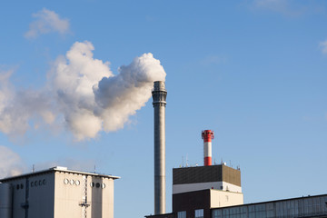 Industrial chimney with smoke and factory buildings against the blue sky