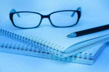 black pen and glasses lying on notebook close up blue color