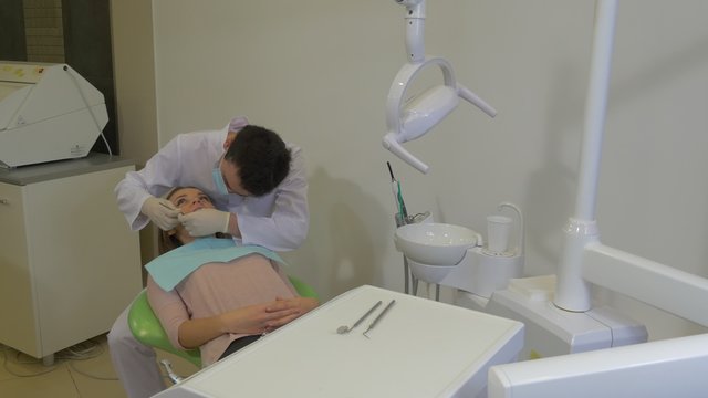 Young Dentist Takes a Tools Examining a Teeth Dentist in Mask is Sitting Behind a Client Woman and Working Concentratedly at Dental Treatment Room