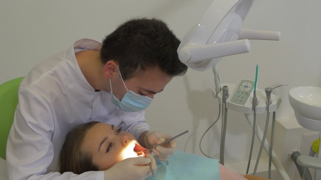Dentist in Mask is Sitting Upon a Patient Examining a Teeth of a Patient by a Mouth Mirror at Dental Treatment Room Green Patient's Chairs Doctor's Chair