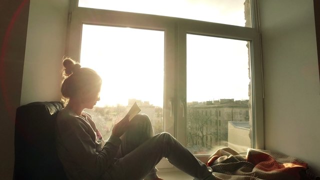 beautiful young woman sitting on window sill with coffee end reading book on background of sunset