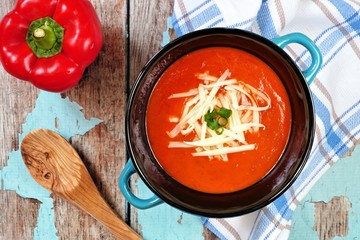 Red pepper soup topped with shredded cheese and green onions, overhead scene on rustic blue wood background