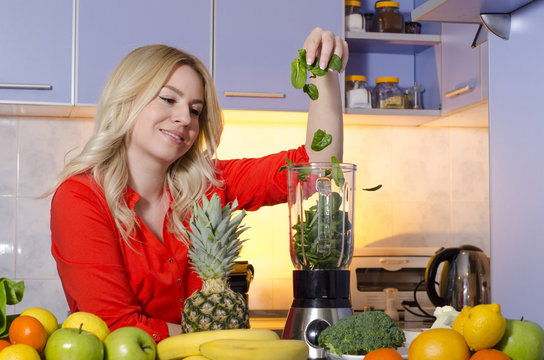 Beautiful young blonde making smoothie from baby spinach and fruits, spinach falling from hands in blender 