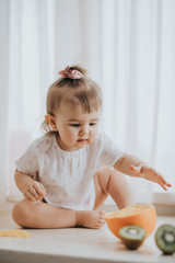 Cute little baby girl on the table in the kitchen eats citrus fruits full of vitamins and makes funny cute faces