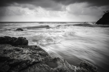 black and white image wave flow on the sandy and rocky beach with dark and dramatic cloud.