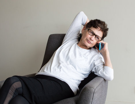 Older Woman With White Top And Glasses Using Smart Phone And Leaning Back In Chair