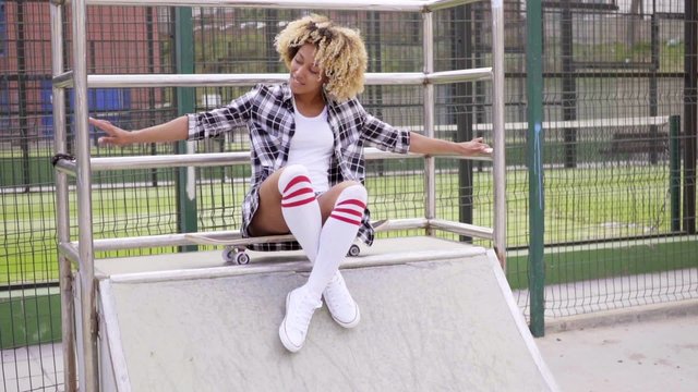 Fun pretty young woman with an afro hairstyle posing on a skateboard on top of a small ramp grinning at the camera