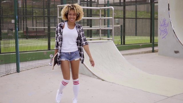 Pretty sporty young woman at a skating rink or park standing smiling at the camera while holding her skateboard behind her back