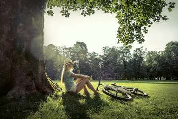 Woman sitting under sun light at day near her bicycle in the par
