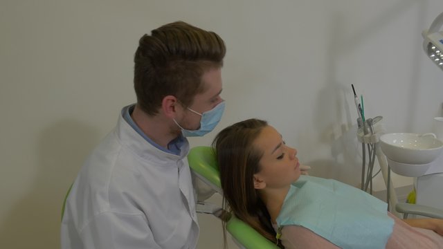 Dentist is Sitting Behind a Client's Head Talking Man in Mask is Examining a Teeth of a Patient Woman With Napkin on Her Chest Dental Clinic Visit