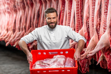 Butcher holding red box full with raw pork standing in the refrigerator with carcasses on the background at the meat manufacturing
