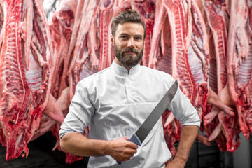 Portrait of a handsome butcher holding knife standing on the pork carcasses background at the meat...