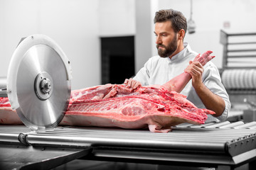 Handsome butcher cutting pork carcasse with big electric saw at the meat manufacturing