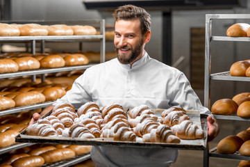 Handsome baker in uniform holding tray full of freshly baked croissants at the manufacturing - obrazy, fototapety, plakaty