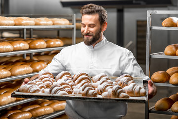 Handsome baker in uniform holding tray full of freshly baked croissants at the manufacturing - obrazy, fototapety, plakaty