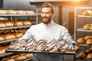 Foto op Plexiglas Knappe bakker in uniform met dienblad vol versgebakken croissants bij de productie © rh2010
