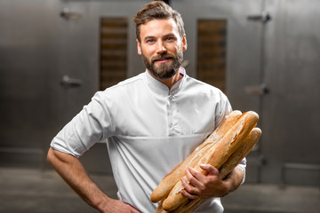 Handsome baker in uniform holding baguettes with oven on the background at the manufacturing