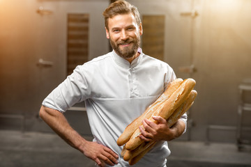 Handsome baker in uniform holding baguettes with oven on the background at the manufacturing - obrazy, fototapety, plakaty