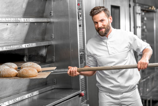 Handsome baker in uniform taking out with shovel freshly baked buckweat bread from the oven at the manufacturing