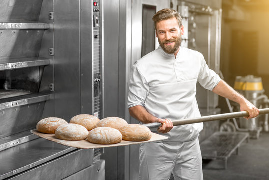 Handsome baker in uniform taking out with shovel freshly baked buckweat bread from the oven at the manufacturing