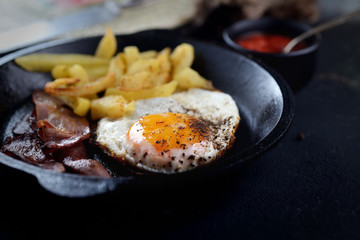 fried eggs with bacon, fried potatoes in a frying pan on a dark background
