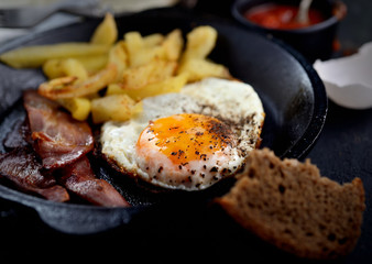 fried eggs with bacon, fried potatoes in a frying pan on a dark background
