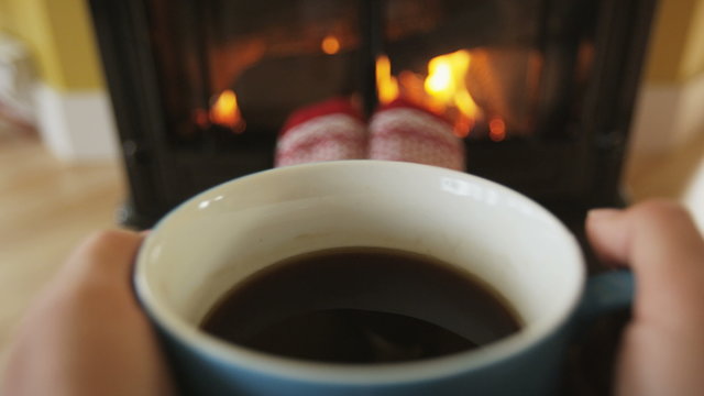 Woman drinking coffee by fireplace getting warm and cozy. Person is having refreshment during in living room. She is warming herself in front of burning stove during winter. RED EPIC SLOW MOTION.