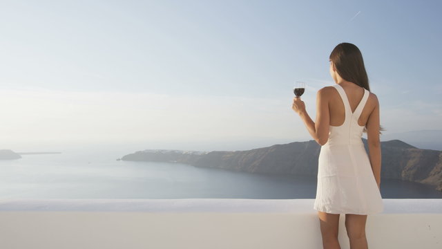 Serenity and relaxation luxury travel lifestyle concept with happy young woman holding red wine glass while standing outside on terrace living jet set lifestyle. Elegant woman, Santorini, Greece.