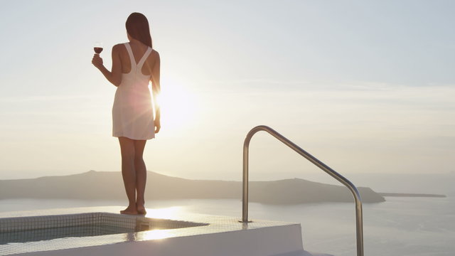 Asian woman drinking red wine while standing at the edge of swimming pool. She is enjoying the view of sea during sunset. Female is wearing sundress during vacation. Santorini, Greece.